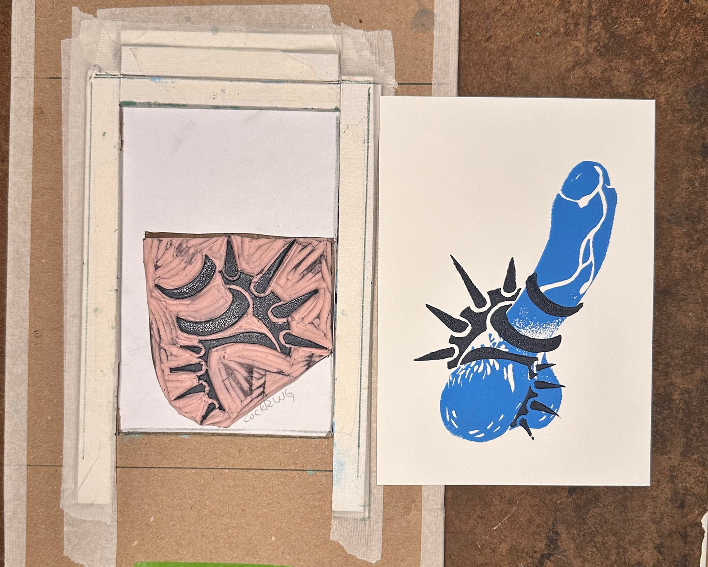 A handmade art print or linocut print sample taped to a workbench under a metal clip. The setup includes a cardboard print block, white tape, a partially colored pink-and-black shield motif, and a separate white print with a blue and black abstract design.